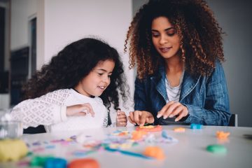 A mom and her daughter sit at a table together playing with small colorful toys. The daughter has a pink sweater and the mom has on a denim blazer.