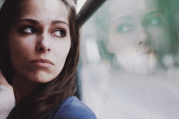 A woman with brown hair and a blue-grey top gazes out a hazy window and is contemplative about life.