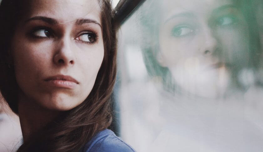 A woman with brown hair and a blue-grey top gazes out a hazy window and is contemplative about life.