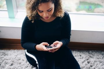 A woman with curly brown hair and a black outfit sits in the window of her high-rise apartment complex. She smiles as she works on her budgeting app on her phone.