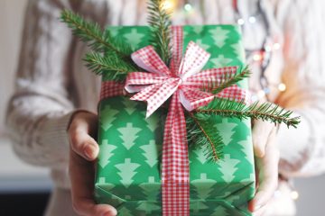 A woman holds a present in green box with a red plaid bow. The green box has trees on it, and her sweater is white. With affordable presents, you can find out how to save money during the holidays.