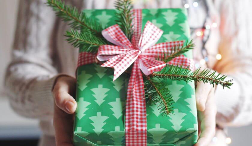 A woman holds a present in green box with a red plaid bow. The green box has trees on it, and her sweater is white. With affordable presents, you can find out how to save money during the holidays.