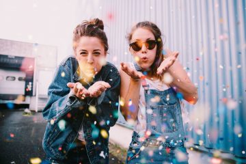 Two friends dressed in blue blow glitter and confetti at the camera. This article explores how to make saving money fun.