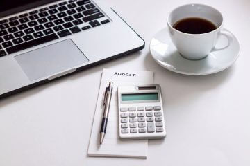 A laptop, coffee cup, and calculator sit on a desk; everything you need to get your budget in order.