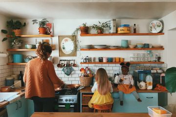 A mother stands at the stove in a kitchen. Two young girls are to her right.