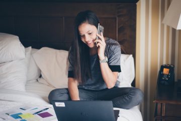 A woman talking on her cell phone in a hotel room as she puts together a new resume. She smiles as she looks at a laptop and paperwork