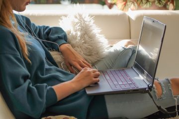 A woman in a dark blue sweatshirt sits on her couch, with a laptop in her lap.