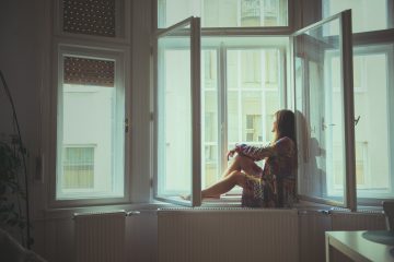 A woman sits at a window and stares at the outside. She appears to be sad.
