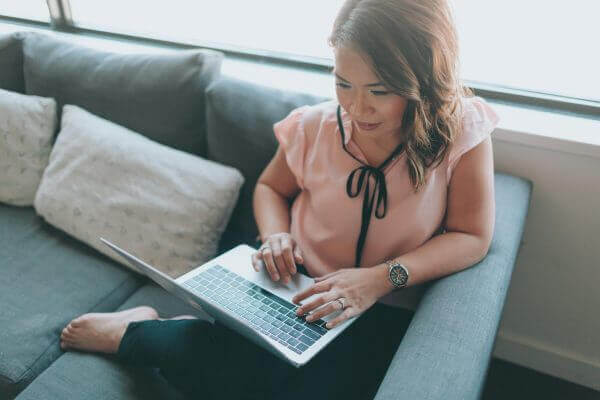 A woman sits on a couch, typing on her laptop. She is wearing a short sleeved shirt and her hair is down.