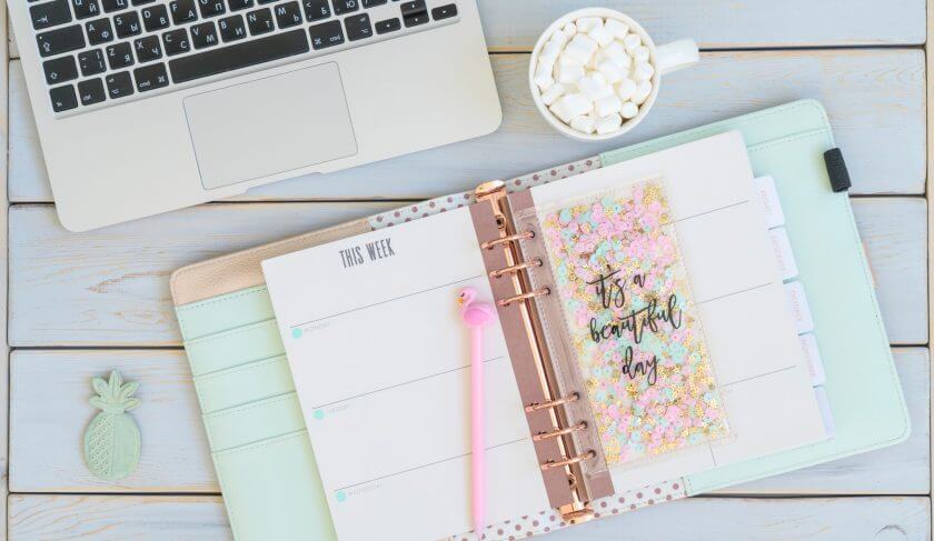 A pastel green and pink planner sits open on a desk next to a cup of coffee and a laptop.