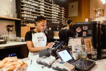 A woman works as a barista at a coffee shop. She wears an apron and stands at a register.
