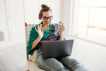 A woman sits at a laptop. She looks excited. She has brown hair and is wearing glasses.