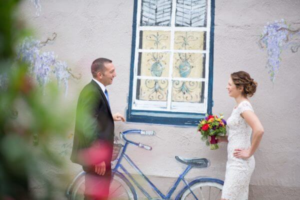 A couple looks at each other on their wedding day. They are standing outdoors in front of a window and bike.