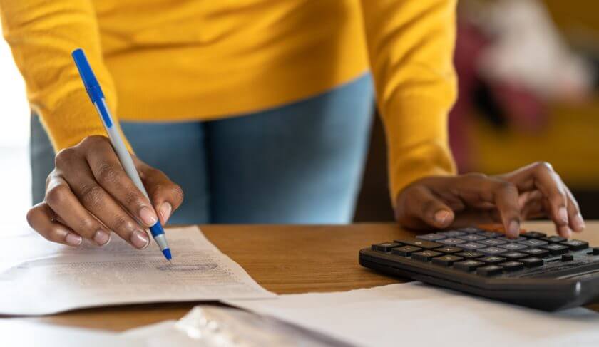 A woman wearing light blue denim jeans and a yellow long-sleeve blouse does taxes at the table, with paperwork, a pen, and a calculator