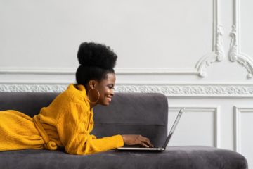 A woman wearing a bright yellow sweater lays on a grey couch and has her laptop open as she types.