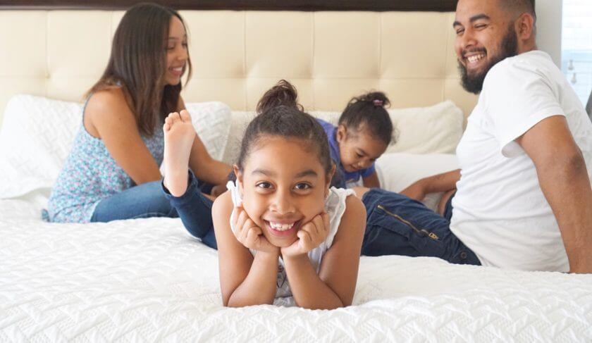 A smiling family of four sit on a bed with a white comforter and chat. A little girl offers a big smile in the center of the bed, with her family behind her.