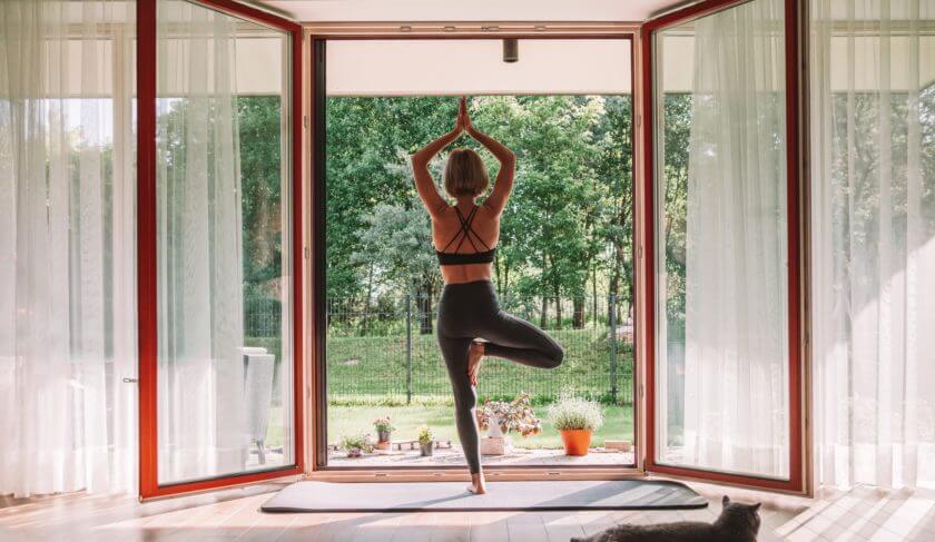 Woman doing yoga on a sunny morning, looking out glass doors at a green yard. She is in black yoga pants, in tree pose.