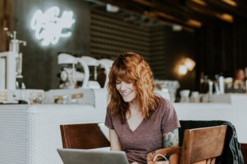 A woman sits in a cafe and works on a laptop. She has red hair and a brown shirt, and smiles as she works.