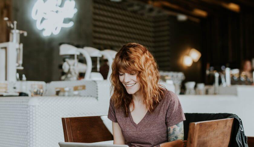 A woman sits in a cafe and works on a laptop. She has red hair and a brown shirt, and smiles as she works.
