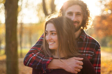 A man and woman stand together. The man is behind the woman, with his arm around her. He has long, curly dark hair and a beard. She has long brown hair. Both are smiling.