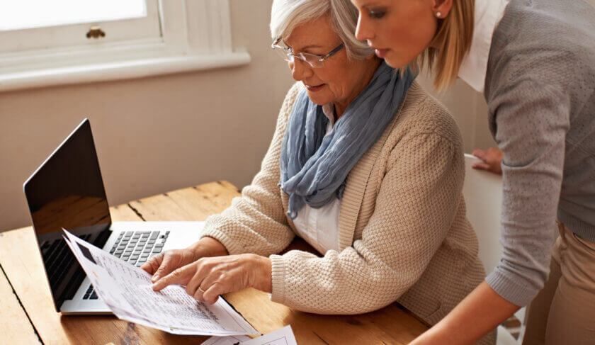 An elderly woman sits at a desk looking at a laptop and paperwork. It appears as though she is being assisted by a younger woman.