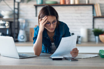 A woman in a blue top leans over the kitchen counter and looks at her budget for the month.
