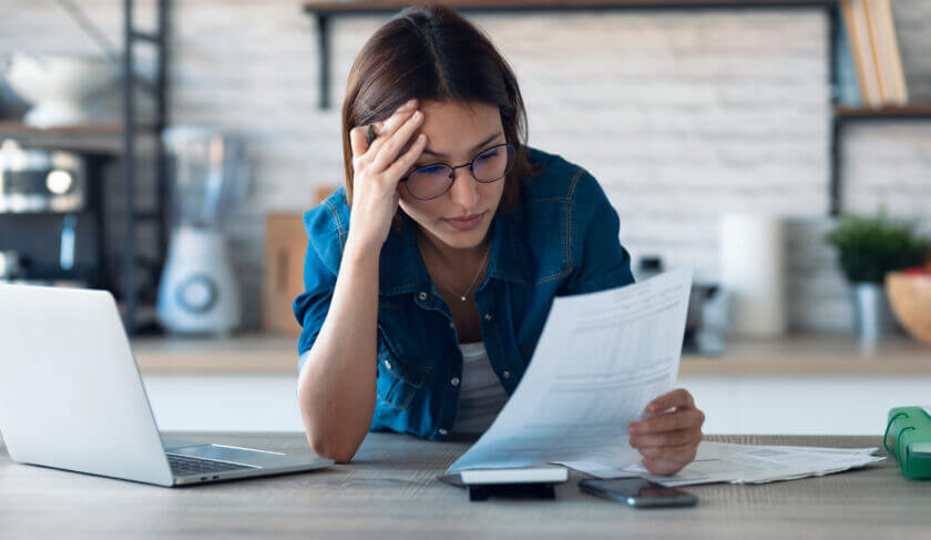 A woman in a blue top leans over the kitchen counter and looks at her budget for the month.