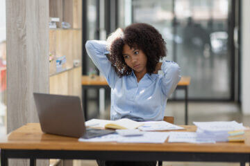 A woman wearing a blue shirt sits at a desk. She looks at her computer and a pile of papers, seeming stressed.
