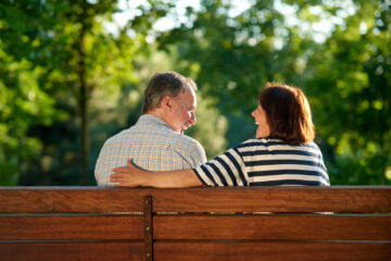 A retired couple sits on a park bench in the sunshine.