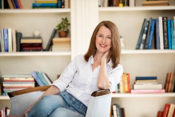 Gretchen Rubin relaxes in a library wearing a white button down shirt and jeans. We discus what it means to reconnect to yourself and your senses