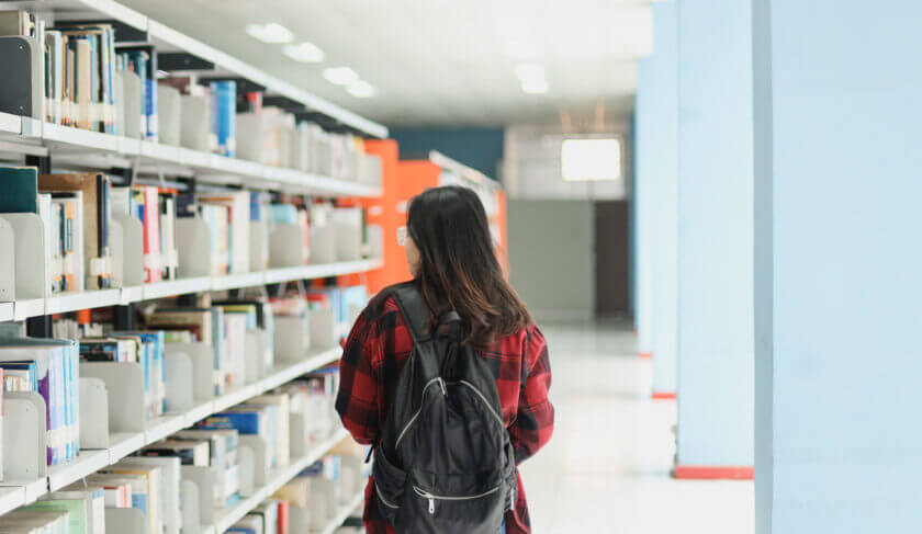 A female college student browses the library on campus, walking between the stacks with a backpack on.