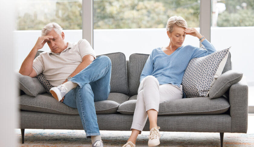 Mature woman and man sit on opposite sides of a couch, looking sad and frustrated.