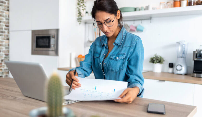 Woman wearing a blue blouse and sitting in her kitchen reviewing financial documents and working on a laptop.