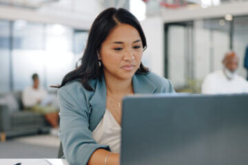 A woman works at her laptop in a bright and open office space. She has dark hair and is wearing a denim blazer.