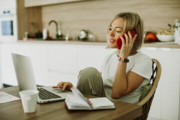 Blonde woman working from home, sitting in her kitchen and chatting on the phone while she works on her laptop.