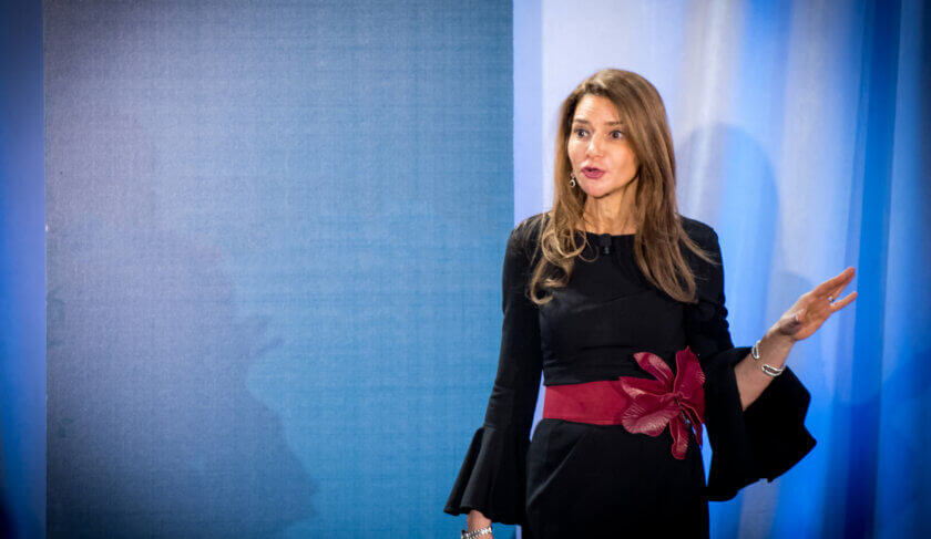 Liz Elting wears a black dress with a cinched red belt giving a speech at a conference.