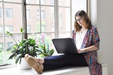 A woman wearing a purple cardigan sits in a sunny window surrounded by plants and works on a laptop.