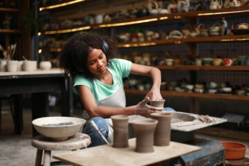 A woman sculpts pottery cups and vases in her pottery studio, where colorful handmade pottery objects line the walls