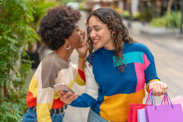 Two women, one carrying shopping bags and wearing colorful sweaters, talk with each other.