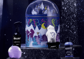 A music box with snow covered mountains on it is pictured in front of a dark backdrop. Bath products are placed to its left and right.