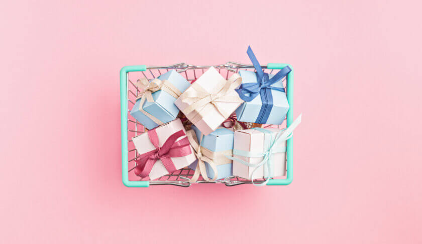 A basket of several holiday gift boxes are pictured on a pink background