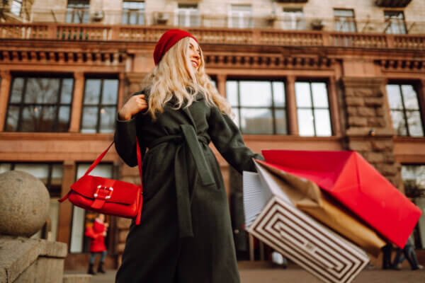 A woman with a red beret and black coat shops along New York City's Fifth Avenue. She carries a red purse and several shopping bags.