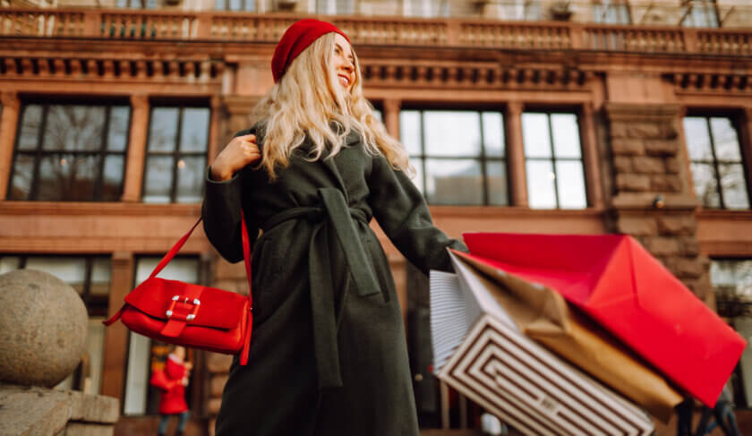 A woman with a red beret and black coat shops along New York City's Fifth Avenue. She carries a red purse and several shopping bags.