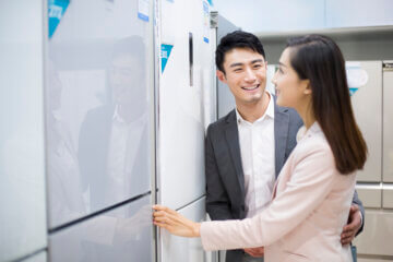 A man and a woman shop for refrigerators at an electronics store. They smile and look at ones that are white and grey in color.