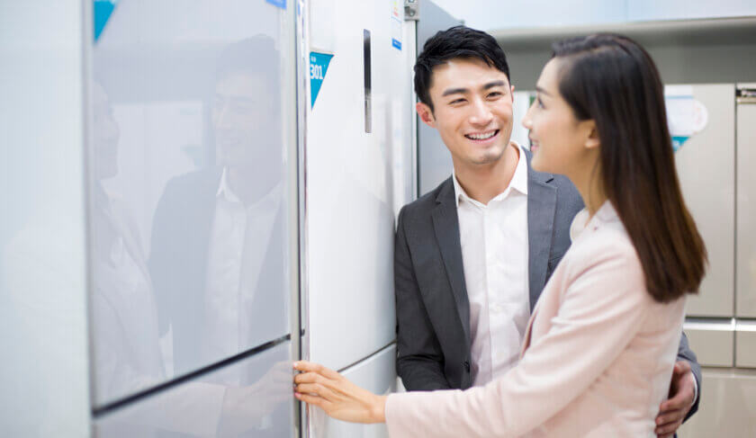 A man and a woman shop for refrigerators at an electronics store. They smile and look at ones that are white and grey in color.