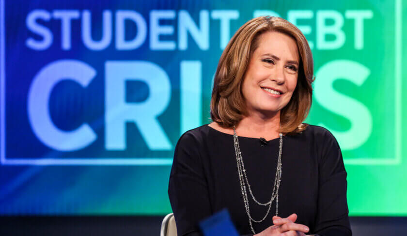 Sheila Bair sits in front of a sign that says 'Student Debt Crisis' and smiles.