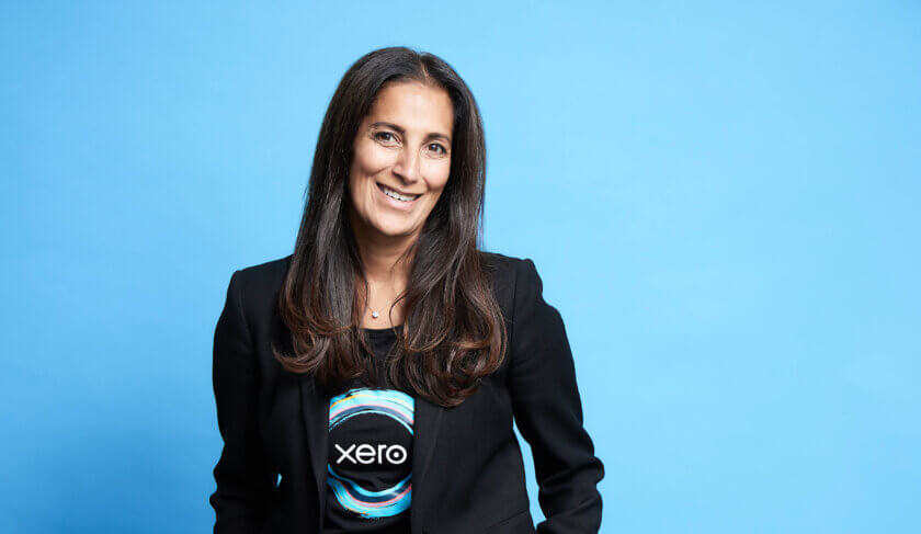 Sukhinder Singh Cassidy stands against a blue background wearing a Xero shirt and smiling at the camera.