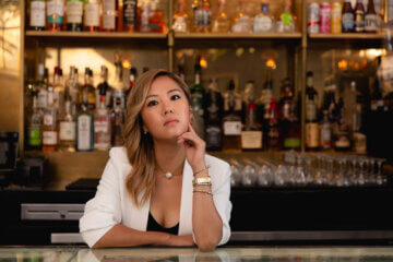 Esther Choi stands behind a bar in a restaurant wearing a white blazer and looking at the camera.