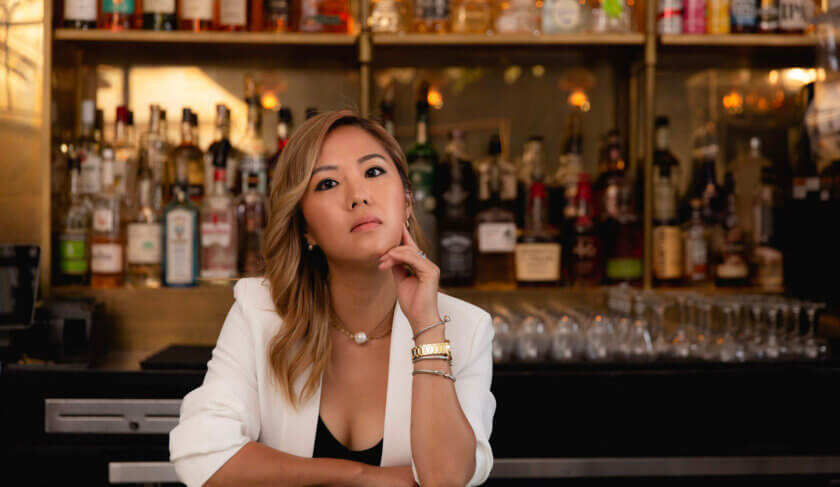 Esther Choi stands behind a bar in a restaurant wearing a white blazer and looking at the camera.