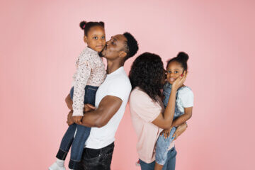A happy family dressed in Jeans and white and pink t-shirts smiles and kisses one another.